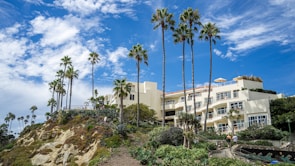A luxurious hillside building with cream-colored walls is surrounded by lush greenery and tall palm trees. The sky above is bright blue with scattered white clouds, and people are walking along a landscaped pathway leading up to the building.