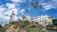A luxurious hillside building with cream-colored walls is surrounded by lush greenery and tall palm trees. The sky above is bright blue with scattered white clouds, and people are walking along a landscaped pathway leading up to the building.