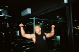 A person with muscular arms is flexing in a gym setting. The individual has short blonde hair and is wearing a sleeveless black shirt with a logo. The background includes gym equipment and dim lighting, contributing to a focused atmosphere.