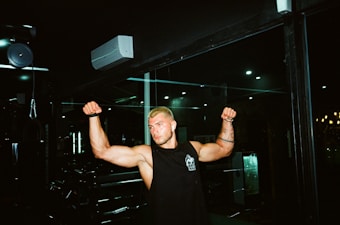 A person with muscular arms is flexing in a gym setting. The individual has short blonde hair and is wearing a sleeveless black shirt with a logo. The background includes gym equipment and dim lighting, contributing to a focused atmosphere.
