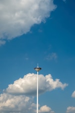 A tall observation tower with a circular viewing platform set against a backdrop of a bright blue sky filled with fluffy white clouds. The tower's sleek design contrasts with the soft shapes of the clouds.