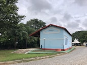 A light blue building with a red roof sits on a cobblestone pathway surrounded by lush green trees and a cloudy sky. The building has the name 'Lagoa da Prata' displayed on its facade. A tent and a few street lamps are also visible.