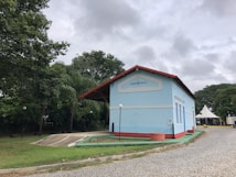 A light blue building with a red roof sits on a cobblestone pathway surrounded by lush green trees and a cloudy sky. The building has the name 'Lagoa da Prata' displayed on its facade. A tent and a few street lamps are also visible.