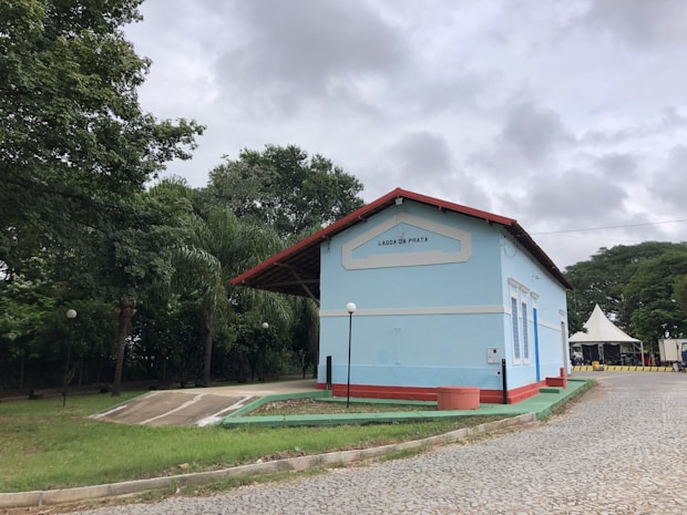 A light blue building with a red roof sits on a cobblestone pathway surrounded by lush green trees and a cloudy sky. The building has the name 'Lagoa da Prata' displayed on its facade. A tent and a few street lamps are also visible.