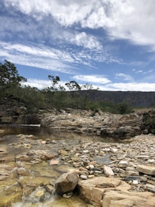 A natural landscape featuring a rocky riverbed with clear water flowing over smooth stones. The background is lined with green trees and shrubbery, set against a mountainside with a partly cloudy blue sky above.