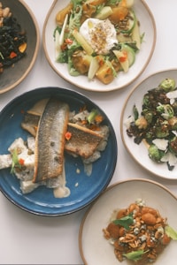 A collection of five dishes is artfully presented on a table. There is a diverse range of foods, including a salad with lettuce, apples, and nuts topped with a creamy dressing, a plate with sautéed fish on a blue plate, roasted Brussels sprouts garnished with cheese, a dish with a vibrant mix of black beans and vegetables, and a bowl of gnocchi topped with sauce and herbs.