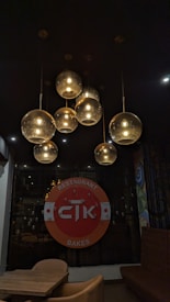 A dimly lit interior features a cluster of hanging spherical pendant lights emitting a warm glow. Below them is a circular sign with the logo 'CTK' and the words 'Restaurant Bakes' surrounded by a decorative pattern. The furnishings include a small wooden table with modern chairs and cushioned seating. The overall ambiance suggests a cozy and intimate dining space.