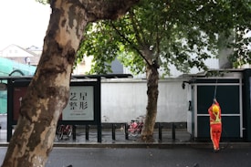 A person in a bright orange uniform and yellow hard hat is power washing the exterior of a structure next to a sidewalk. Large trees with dense green foliage are visible, along with bicycles parked under a canopy. The scene appears to be in an urban setting with buildings and construction in the background.