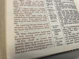 The image displays an open page of a Bible featuring text in both standard black font and highlighted red font. The page contains biblical verses with numerical references and footnotes on the side providing additional information. The physical book appears somewhat worn, with visible edges showing signs of use.