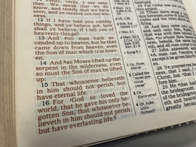 The image displays an open page of a Bible featuring text in both standard black font and highlighted red font. The page contains biblical verses with numerical references and footnotes on the side providing additional information. The physical book appears somewhat worn, with visible edges showing signs of use.