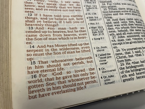 The image displays an open page of a Bible featuring text in both standard black font and highlighted red font. The page contains biblical verses with numerical references and footnotes on the side providing additional information. The physical book appears somewhat worn, with visible edges showing signs of use.