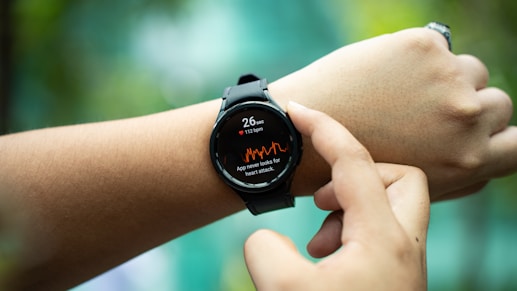 A smartwatch on a person's wrist displays heart rate information and a warning about potential heart attack detection. The watch has a black strap and the display shows a heart rate of 112 bpm with red waveform graphics. Another hand is pointing to or interacting with the watch.