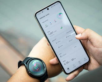 A person holds a smartphone displaying the Samsung Health app, showing metrics such as daily activity, steps, calories, and exercise data. The individual also wears a smartwatch on their wrist, which has a colorful display indicating health statistics.