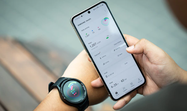 A person holds a smartphone displaying the Samsung Health app, showing metrics such as daily activity, steps, calories, and exercise data. The individual also wears a smartwatch on their wrist, which has a colorful display indicating health statistics.