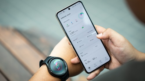 A person holds a smartphone displaying the Samsung Health app, showing metrics such as daily activity, steps, calories, and exercise data. The individual also wears a smartwatch on their wrist, which has a colorful display indicating health statistics.