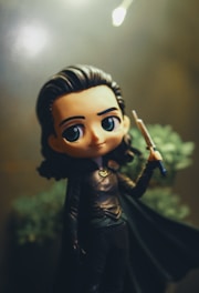 A chibi-style figurine is depicted with large, expressive eyes and long dark hair, dressed in a dark outfit with a cape. The figure holds a short sword, and the background is blurred with soft light, adding a mystical ambiance.
