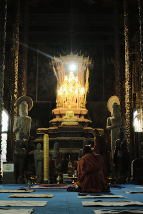 A serene temple interior featuring a large altar with a golden Buddha statue. Monks in orange robes are engaged in prayer on a blue carpet, surrounded by other Buddha statues. The setting is dimly lit except for the glowing altar and ornate decorations.
