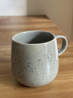 A ceramic mug with a speckled pattern sits on a wooden surface. The mug is light gray with small, irregular dark spots, and has a round shape with a handle. Soft natural light highlights its texture, casting a gentle shadow on the wood.