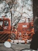 Firefighters in orange uniforms are gathered around a fire truck. They appear to be on standby, taking a break with some sitting on the ground and others leaning against the truck. The scene is shaded by tall trees, giving a calm atmosphere despite the emergency vehicle present.