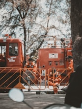 Firefighters in orange uniforms are gathered around a fire truck. They appear to be on standby, taking a break with some sitting on the ground and others leaning against the truck. The scene is shaded by tall trees, giving a calm atmosphere despite the emergency vehicle present.