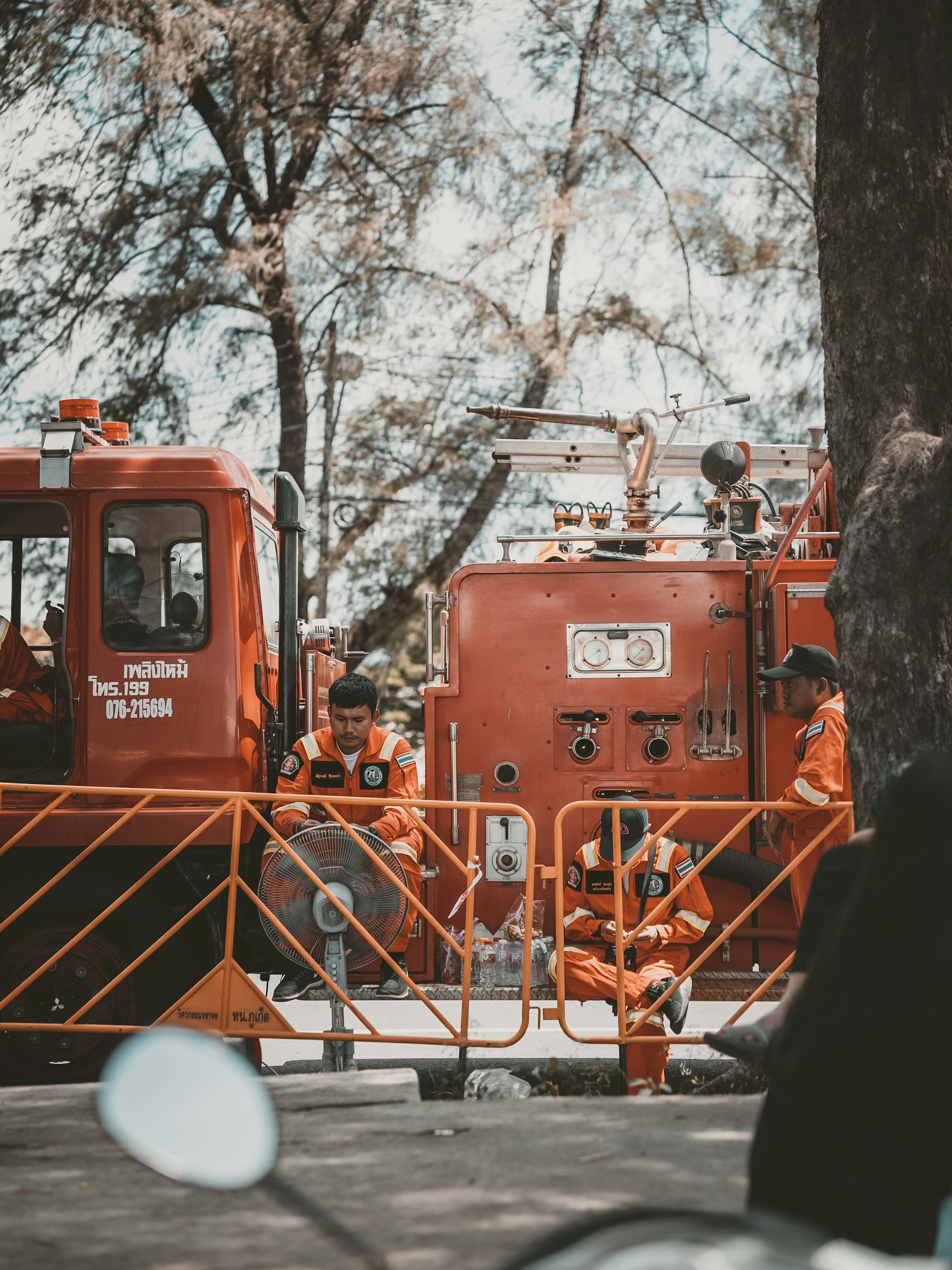 Firefighters in orange uniforms are gathered around a fire truck. They appear to be on standby, taking a break with some sitting on the ground and others leaning against the truck. The scene is shaded by tall trees, giving a calm atmosphere despite the emergency vehicle present.