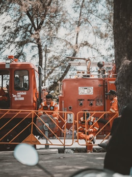 Firefighters in orange uniforms are gathered around a fire truck. They appear to be on standby, taking a break with some sitting on the ground and others leaning against the truck. The scene is shaded by tall trees, giving a calm atmosphere despite the emergency vehicle present.
