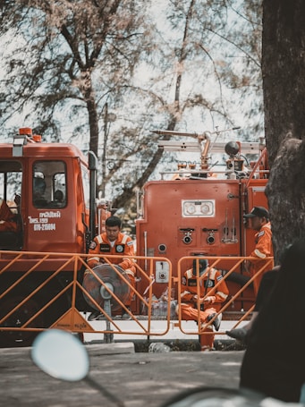 Firefighters in orange uniforms are gathered around a fire truck. They appear to be on standby, taking a break with some sitting on the ground and others leaning against the truck. The scene is shaded by tall trees, giving a calm atmosphere despite the emergency vehicle present.