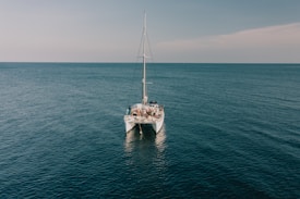 A catamaran is sailing on a vast, calm ocean with a group of people on its deck. The sea appears tranquil under a clear sky with a hint of clouds towards the horizon.