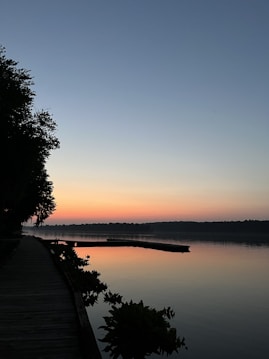 A serene lakeside scene during sunset or sunrise, with calm water reflecting the sky's gradient, transitioning from pale orange near the horizon to a soft blue higher up. A wooden deck runs along the left side, bordered by vegetation, and extends towards the water with trees lining the shoreline.