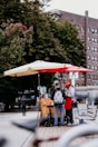 A street vendor wearing a red apron and cap serves a customer under large umbrellas. The vendor cart is decorated with floral patterns and surrounded by various items, including bottled beverages. The setting is urban, with trees and a large brick building in the background.