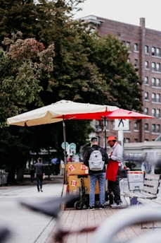 A street vendor wearing a red apron and cap serves a customer under large umbrellas. The vendor cart is decorated with floral patterns and surrounded by various items, including bottled beverages. The setting is urban, with trees and a large brick building in the background.
