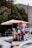 A street vendor wearing a red apron and cap serves a customer under large umbrellas. The vendor cart is decorated with floral patterns and surrounded by various items, including bottled beverages. The setting is urban, with trees and a large brick building in the background.