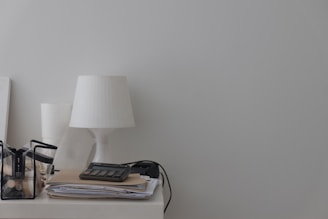 A minimalist desk setup with a white lamp, a transparent container holding various stationary items, a calculator on top of a stack of papers, and a power cable visible.