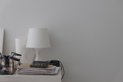 A minimalist desk setup with a white lamp, a transparent container holding various stationary items, a calculator on top of a stack of papers, and a power cable visible.