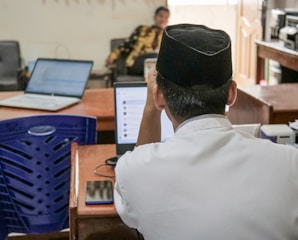 A person wearing a traditional hat is seated at a desk with a laptop open, surrounded by other office equipment. A blue plastic chair is nearby, and another person is seen in the background sitting on a chair.
