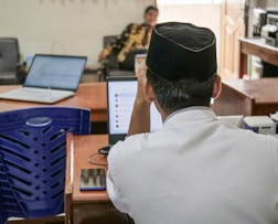 A person wearing a traditional hat is seated at a desk with a laptop open, surrounded by other office equipment. A blue plastic chair is nearby, and another person is seen in the background sitting on a chair.