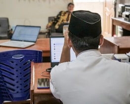 A person wearing a traditional hat is seated at a desk with a laptop open, surrounded by other office equipment. A blue plastic chair is nearby, and another person is seen in the background sitting on a chair.