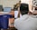 A person wearing a traditional hat is seated at a desk with a laptop open, surrounded by other office equipment. A blue plastic chair is nearby, and another person is seen in the background sitting on a chair.