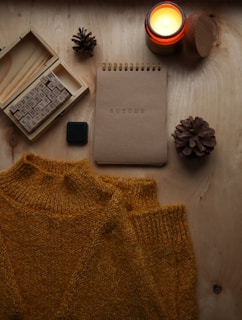 A wooden surface is adorned with a cozy, mustard-colored sweater, an open box of alphabet stamps, a burning candle in a small jar, a notebook with 'AUTUMN' embossed on its cover, and two pinecones. The items are arranged in an aesthetically pleasing manner, evoking a sense of warmth and comfort.