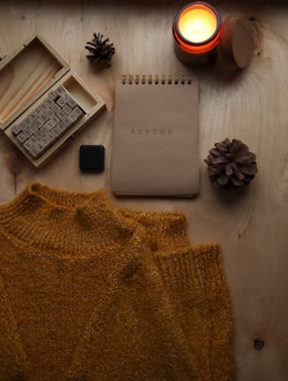 A wooden surface is adorned with a cozy, mustard-colored sweater, an open box of alphabet stamps, a burning candle in a small jar, a notebook with 'AUTUMN' embossed on its cover, and two pinecones. The items are arranged in an aesthetically pleasing manner, evoking a sense of warmth and comfort.