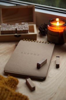 A cozy setting featuring a candle burning warmly beside an open box of alphabet stamps. A notebook labeled 'AUTUMN' is placed on a wooden surface along with scattered stamps. A knitted fabric partially visible adds a touch of warmth to the scene.