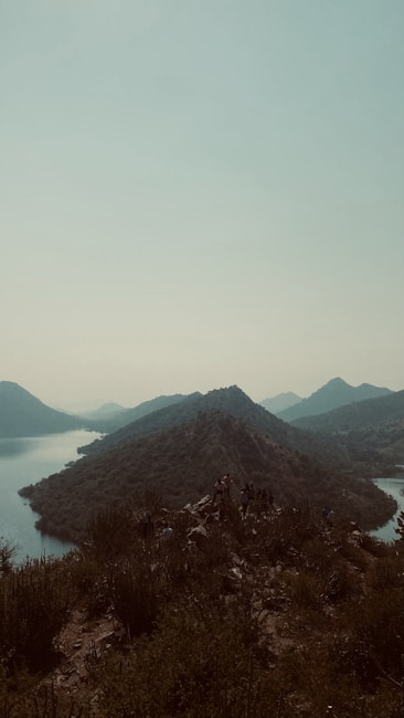A tranquil landscape featuring a series of small, lushly vegetated hills surrounded by calm water bodies under a pale, cloudy sky. A group of people can be seen congregated on one of the hills in the foreground, adding a sense of exploration or adventure to the scene.