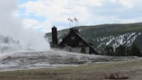 A rustic lodge with multiple flagpoles is set against a backdrop of a forested hillside. Steam is rising from a large geyser in the foreground, with cloudy skies and partially snow-covered trees in the background.