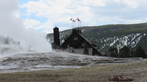 A rustic lodge with multiple flagpoles is set against a backdrop of a forested hillside. Steam is rising from a large geyser in the foreground, with cloudy skies and partially snow-covered trees in the background.