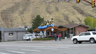 A building with a sculpture of people in a blue raft is visible on the roof. The scene includes a street with cars and pedestrians. A mountain ridge forms the backdrop, and there are trees and a clear sky. The building has large glass windows and a wooden awning.