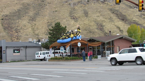 A building with a sculpture of people in a blue raft is visible on the roof. The scene includes a street with cars and pedestrians. A mountain ridge forms the backdrop, and there are trees and a clear sky. The building has large glass windows and a wooden awning.