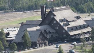 A large, rustic lodge with multiple steep roofs and dark wooden exterior sits in a forested area. Several people are visible near the entrance, and a few cars are parked nearby. Trees are surrounding the building, and a flag is visible on top of the structure.