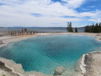 A large, vibrant blue geothermal hot spring surrounded by mineral deposits. A wooden boardwalk runs alongside the hot spring, with a group of people standing and observing the natural feature. Beyond the hot spring, a lake stretches into the distance with a line of trees and distant mountains under a partly cloudy sky.