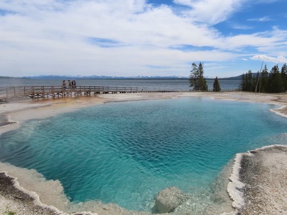 A large, vibrant blue geothermal hot spring surrounded by mineral deposits. A wooden boardwalk runs alongside the hot spring, with a group of people standing and observing the natural feature. Beyond the hot spring, a lake stretches into the distance with a line of trees and distant mountains under a partly cloudy sky.