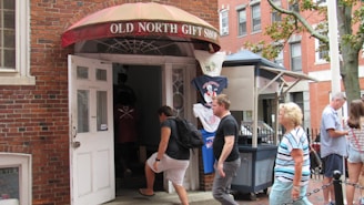 A group of people are walking in and out of a gift shop located in a brick building. The shop is labeled 'Old North Gift Shop' with an awning above the open door. T-shirts are displayed on racks outside the shop, and there is a ticket booth nearby. A tree and additional buildings are visible in the background.